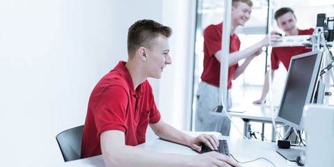 zwei Azubis mit roten T-Shirt an einer Maschine, ein Azubi mit rotem T-Shirt sitzt am Computer
