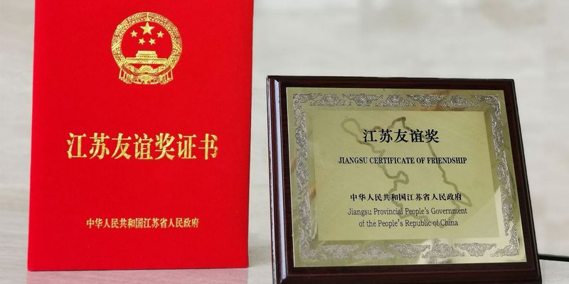 Freundschaftspreise, links rotes kleines Buch mit chinesischer goldener Aufschrift und rechts ein goldenes Bild mit holz Umrandung und schwarzer Schrift "Jiangsu Certificate of Friendship - Jiangsu Provincial People´s Government of the People´s Republic of China"