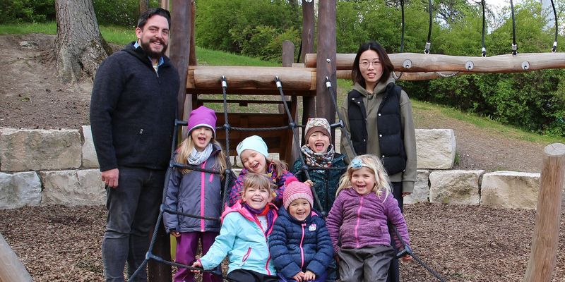 Kindergarten Klettergerüst sechs Kinder sitzen auf seilen zwei Erwachsene Personen daneben ein dunkelhaariger Mann mit Bart eine asiatische Frau mit Brille 