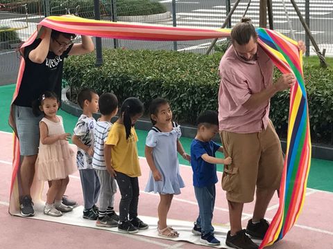 Kinder und Erzieher spielen draußen großes Regenbogenband um die Kinder und die Erzieher auf Sportplatz